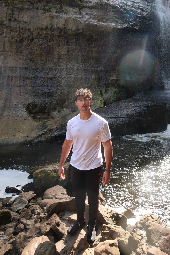 Adam in a white t-shirt standing on rocks at the base of a waterfall, sunlight flaring off the water behind him